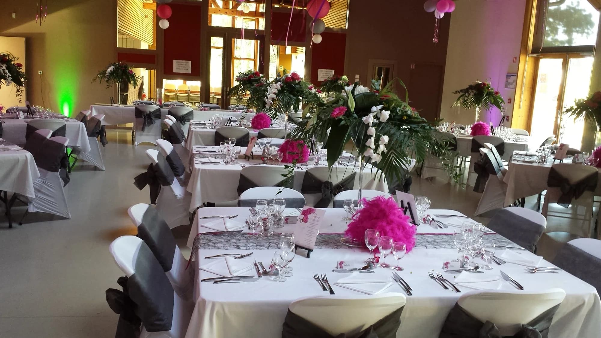 Salle de mariage avec tables rondes dressées, hautes compositions florales blanches et fuchsia et housses de chaises à nœuds gris